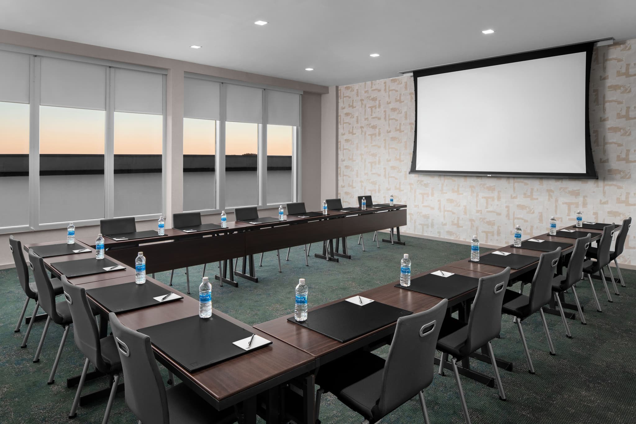 A professional meeting room at AC Hotels Raleigh, set up in a U-shape with black tables, chairs, bottled water, and a large projector screen.