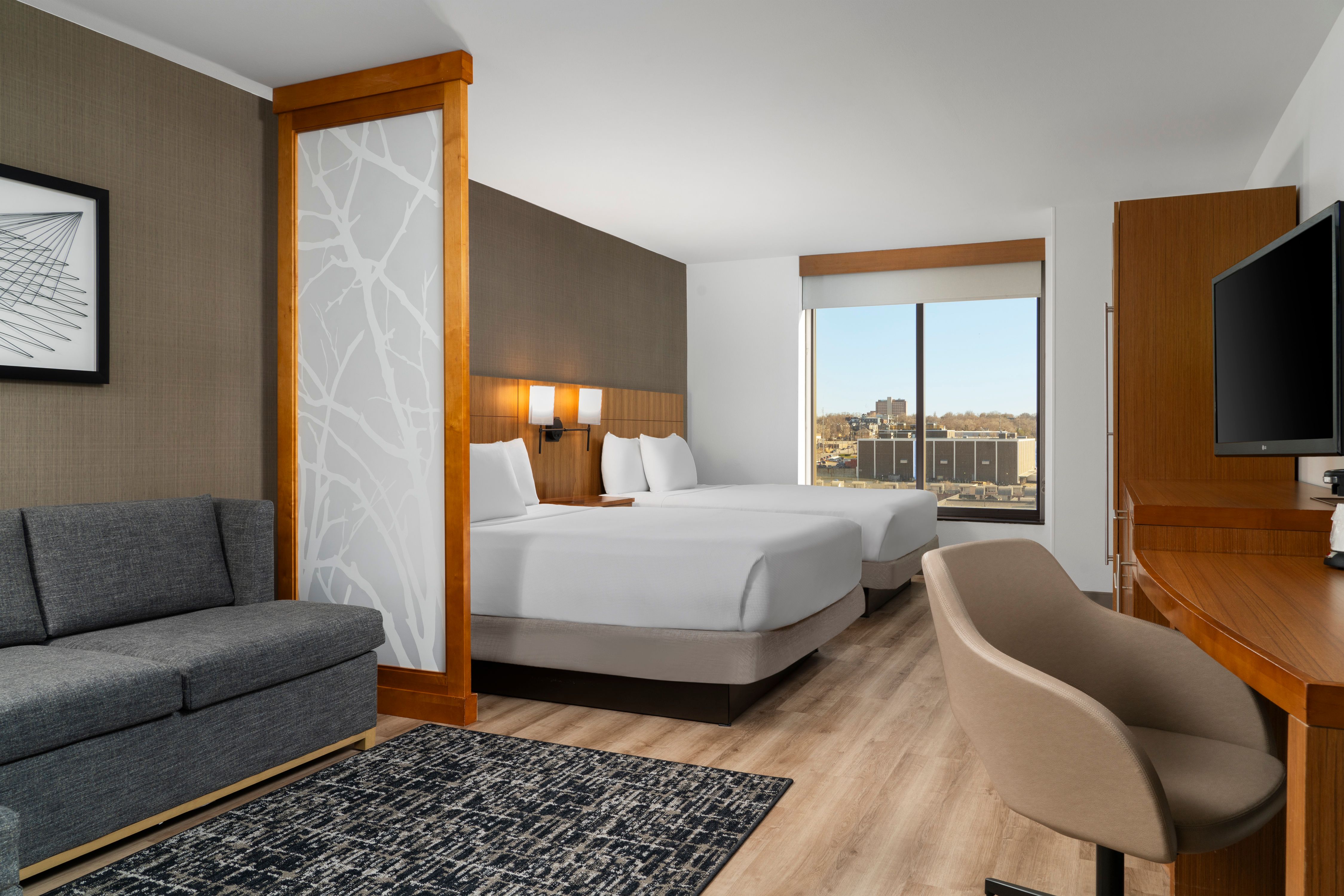 A stylish hotel room at Hyatt Place Omaha, featuring a cozy grey sofa, large windows and 2 single beds.