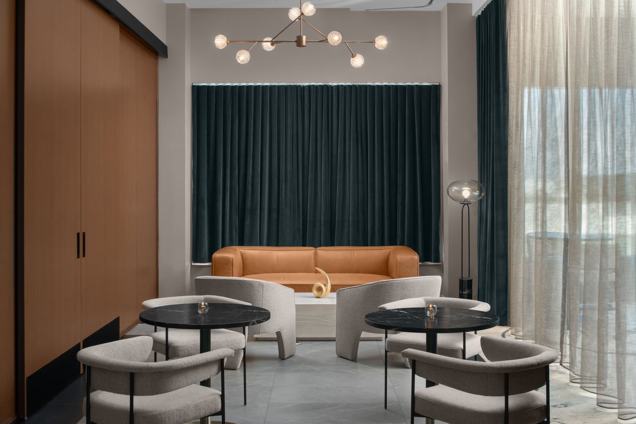 A stylish Level 7 lounge room at AC Hotels Raleigh, featuring a cozy seating area with a tan leather sofa, modern round tables with plush chairs, dark green curtains, and warm ambient lighting.