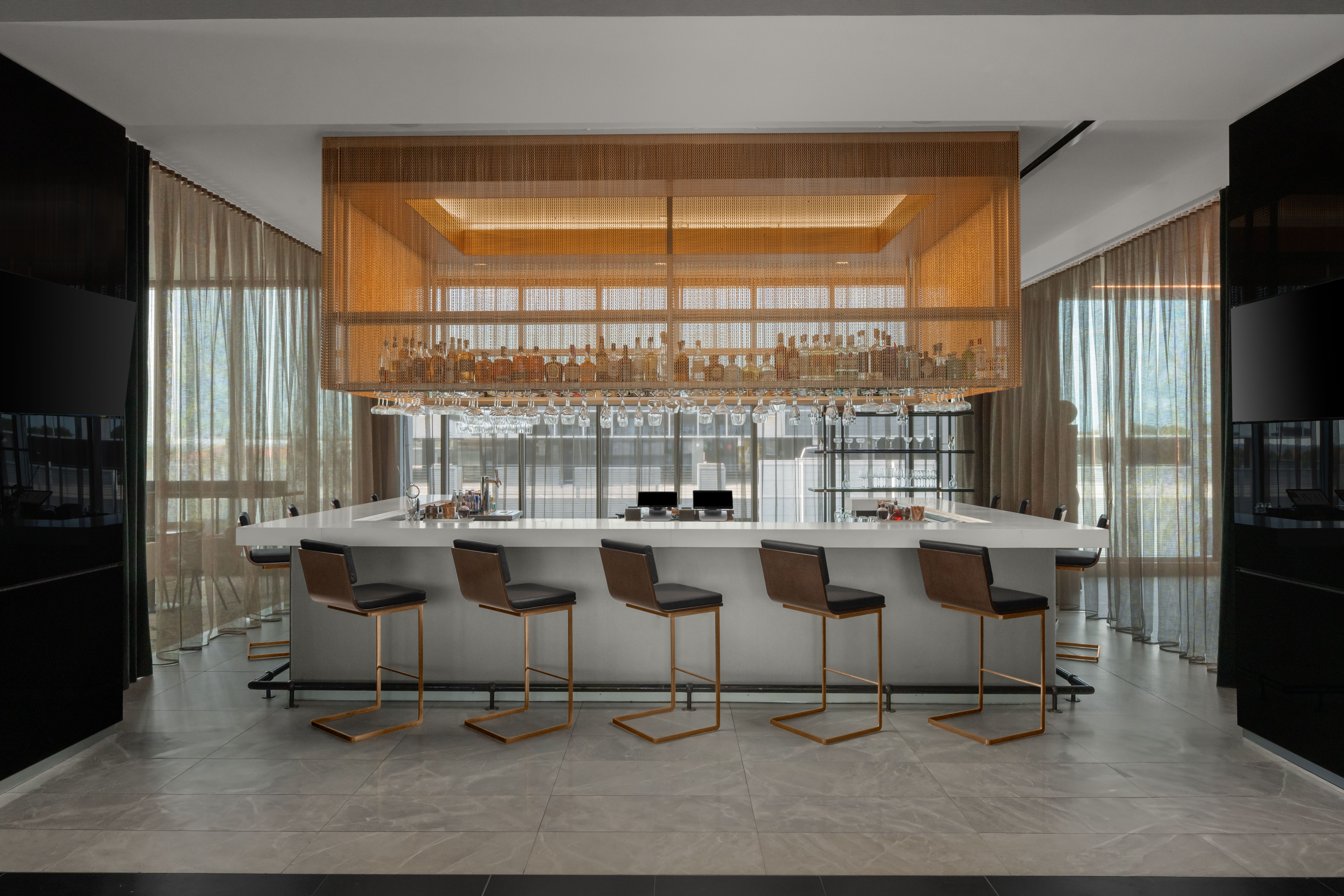 Sophisticated indoor bar at AC Hotels Raleigh, featuring a sleek white counter, modern barstools, and an elegant suspended liquor display.